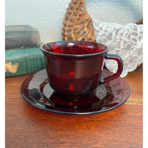 Puede incluir: Un juego de taza de té y platillo a juego de cristal en color rojo rubí intenso. La taza presenta un acabado liso y translúcido con forma de cuenco redondeado y un asa lateral resistente. El platillo es circular con un centro elevado.
