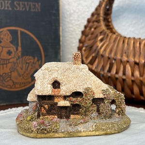 Pode incluir: Uma estatueta de casa em miniatura com um telhado de palha texturizado, uma chaminé de tijolos e paredes de madeira escura. A estrutura é adornada com trepadeiras, pequenas flores cor-de-rosa e uma base de pedra, criando uma cena detalhada.