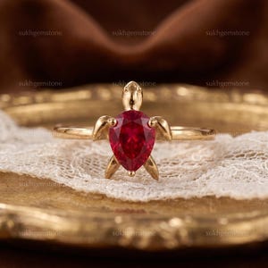 Puede incluir: Un anillo dorado con un diseño de tortuga, engastado con una gema roja en forma de pera. El anillo se exhibe sobre una superficie de encaje blanco. El nombre de la marca "sukhgemstone" es visible en el fondo.