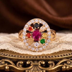 May include: A gold ring with a central oval ruby, encircled by white gemstones and smaller, colourful gems. The ring has a detailed band and is set on a decorative surface. The text "10 SUKHGEMSTONE" is visible.