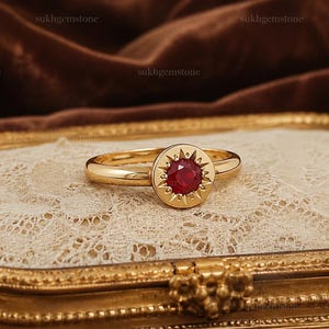Op de afbeelding: Een gouden ring met een ronde, gefacetteerde rode edelsteen in het midden van een ronde plaat met een zonnestraalontwerp. De metalen band is glad en gepolijst, en de zetting is gedetailleerd met kleine, uitstralende stralen rond de steen.