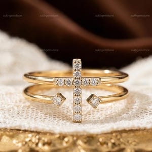 May include: A gold-colored ring featuring a cross design adorned with small, sparkling gemstones. The ring has a double-band design with a gemstone accent on each side. The ring is displayed on a white lace surface.