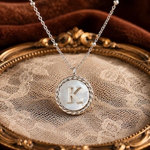 May include: A silver-toned necklace featuring a circular pendant with a white mother-of-pearl inlay and the letter K centred in a serif font. The pendant is framed by a twisted rope-style border and hangs from a delicate chain with small beads.