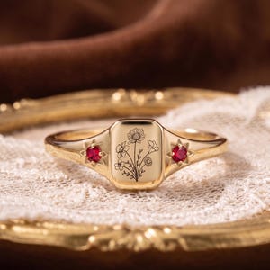 Può includere: Un anello dorato con una faccia rettangolare incisa con fiori. Due piccole pietre rosse rotonde sono incastonate su entrambi i lati della faccia. L'anello è esposto su una superficie di pizzo bianco.
