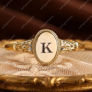 Può includere: Un anello in oro caratterizzato da una parte frontale ovale con bordo perlato e la lettera K incisa al centro. Il gambo presenta un motivo complesso e strutturato a forma di foglia su entrambi i lati, che si estende dalla montatura ovale.