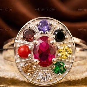 May include: A silver ring featuring a large oval ruby at the center, surrounded by eight smaller gemstones of various colors, including red, purple, black, and yellow. The ring has a detailed, segmented design and is set against a neutral background.
