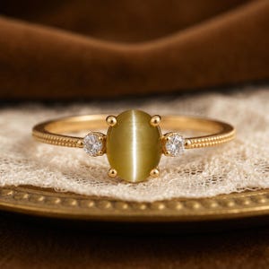 Op de afbeelding: Een gouden ring met een centrale ovale olijfgroene kattenoogsteen, vastgezet met vier pootjes. De ringband is aan weerszijden versierd met een kleine, heldere, rondgeslepen steen en heeft een verfijnde textuur met kleine kraaltjes.