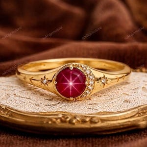 May include: A gold ring with a large, round, red gemstone at the center, displaying a star-like pattern. The ring is adorned with small diamonds and celestial designs. The ring is displayed on a decorative, gold-trimmed tray.