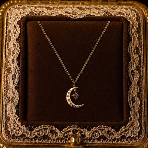 Pode incluir: Um colar de tom dourado com um pingente de meia-lua adornado com pequenas estrelas gravadas e um berloque de estrela pendente com uma pequena pedra preciosa vermelha. A corrente delicada está presa ao topo do pingente de meia-lua.