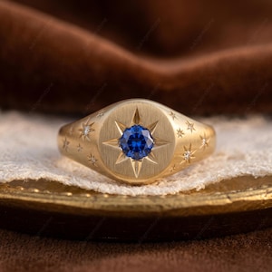 May include: A gold signet ring featuring a central blue gemstone in an eight-pointed star design. The ring is adorned with small star and diamond accents. The ring is displayed on a lace-like surface.