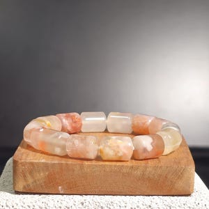 Puede incluir: Un brazalete hecho de cuentas rectangulares translúcidas en tonos naranja, blanco y amarillo. El brazalete se exhibe sobre un pequeño bloque de madera rectangular. El fondo es gris oscuro y el primer plano es una superficie blanca texturizada.