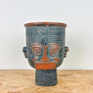 Puede incluir: Un jarrón decorativo de cerámica con diseño de rostro. El jarrón presenta rasgos faciales anaranjados y un cuerpo verde oscuro. El borde es de color marrón rojizo. El jarrón está sobre una superficie de madera.
