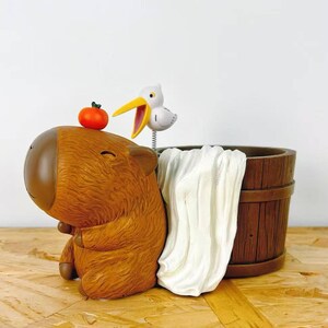 May include: A brown capybara figurine with a small orange tomato on its head, next to a wooden barrel and a white towel. A small bird with a yellow beak is perched above the barrel. The figurine is on a wooden surface.