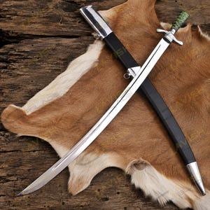 May include: A curved steel sword with a polished blade and a green, textured handle featuring a silver crossguard. The sword is accompanied by a black scabbard with silver metal fittings at the top and bottom, positioned diagonally across the blade.