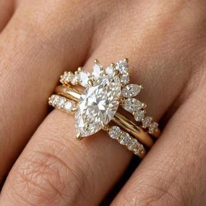 May include: A trio of gold rings set with diamonds. The central ring has a marquise-cut diamond, with smaller diamonds around it. The other two rings are embellished with round diamonds, creating a layered look. The rings are on a hand.