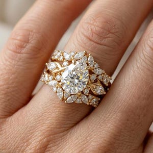 May include: A gold ring featuring a central pear-shaped diamond surrounded by a cluster of smaller marquise and round-cut diamonds. The band is crafted from yellow gold and incorporates a delicate, branching design that frames the center stone.