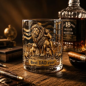 Peut inclure: Un verre à whisky transparent orné d'un relief doré représentant un lion et trois lionceaux dans une savane. Le texte "Best DAD ever" est gravé en lettres dorées à la base. Le décor inclut des lignes ondulées et des silhouettes d'arbres.
