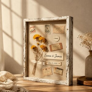 Può includere: Una cornice a cassetta rettangolare con finitura bianca anticata e fondo in lino. All'interno si trovano fiori gialli essiccati, francobolli in stile vintage, etichette di carta e due targhette con la scritta Emma & James e June 15, 2026.