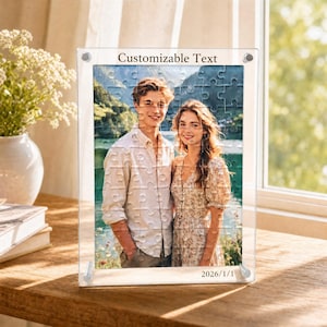 Può includere: Una cornice trasparente in acrilico per puzzle con una fotografia di una coppia. Il puzzle è composto da pezzi ad incastro. Il testo "Customizable Text" è in alto e "2026/1/1" in basso. La cornice è su una superficie di legno.