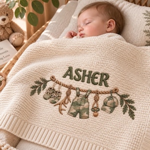 May include: A cream-coloured knitted baby blanket featuring the embroidered name ASHER in dark green. The design includes a rope motif with hanging baby items: booties, a pacifier, overalls with a camouflage pattern, and a matching cap.