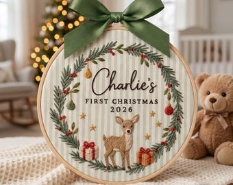 May include: A circular embroidery hoop featuring a light green and white striped fabric background. The design includes a wreath of pine branches, red berries, small ornaments, and gold stars. Centered text reads Charlie's FIRST CHRISTMAS 2026 above a small deer and two gift boxes. A large olive green satin ribbon is tied at the top.