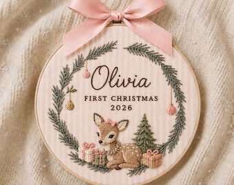 May include: A circular embroidery hoop featuring a light pink and white striped fabric background. The design includes a green pine wreath, a central fawn illustration, and the text Olivia FIRST CHRISTMAS 2026. A pink satin ribbon bow is attached at the top.