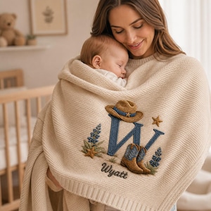 Pode incluir: Uma manta de bebé em malha bege com um design bordado de uma letra W grande em azul, um chapéu de cowboy castanho, um par de botas de cowboy castanhas, flores azuis e uma pequena estrela. O nome Wyatt está bordado em fio escuro abaixo da letra W.