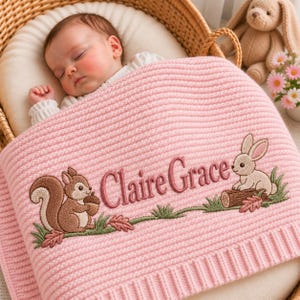 May include: A soft pink knitted baby blanket featuring an embroidered design of a brown squirrel holding an acorn and a light brown rabbit sitting on a log. The name Claire Grace is embroidered in dark pink script between the animals.