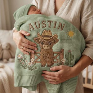 May include: A sage green knitted baby blanket featuring the embroidered name AUSTIN in brown letters. The design includes a brown highland cow wearing a cowboy hat behind a wooden fence, a yellow sun, a green cactus, and white flowers.