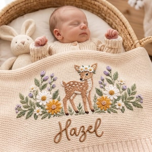 May include: A cream-coloured knit baby blanket featuring an embroidered brown fawn wearing a flower crown, surrounded by white and yellow daisies and purple sprigs. The name Hazel is embroidered in brown script below the fawn, with a plush toy nearby.