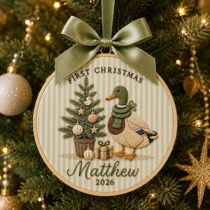 May include: A round embroidery hoop ornament featuring a light green striped fabric background. It displays a stitched Christmas tree, a mallard duck wearing a green scarf, a gift, and the text "FIRST CHRISTMAS Matthew 2026". A sage green satin ribbon bow is attached to the top.
