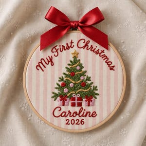 Könnte beinhalten: Ein runder Stickrahmen mit hellrosa und weiß gestreiftem Stoff. Das Motiv zeigt einen grünen Weihnachtsbaum mit roten und rosa Kugeln, drei Geschenken am Fuß sowie den gestickten Text My First Christmas Caroline 2026.