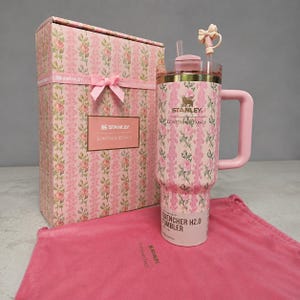 May include: A pink Stanley Quencher H2.0 Tumbler with a floral pattern, a handle, and a straw, next to a matching gift box and a pink drawstring bag. The tumbler and box feature the text "Stanley LovesShackFancy". The gift box is tied with a pink ribbon.