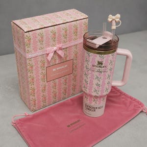 May include: A pink Stanley Quencher H2.0 tumbler with a floral pattern, a matching handle, and a straw. It is presented with a pink gift box and a pink drawstring bag. The box and tumbler feature the text "Stanley LovesShackFancy".