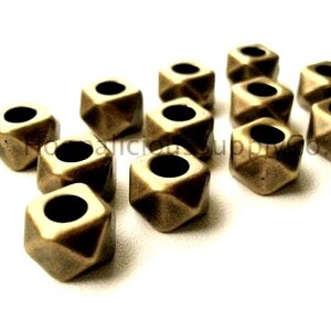 May include: A collection of bronze-colored, geometric beads with a square hole in the center. The beads have a faceted design, creating multiple planes on each side. The beads are scattered on a white surface, showcasing their shape and color.