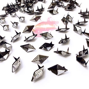 May include: A scattering of silver-toned diamond-shaped metal studs with pointed tips. The studs are scattered on a white surface.