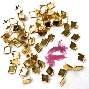 May include: A scattering of gold-coloured metal rivets on a white surface. A pink lipstick mark is visible in the centre of the image.