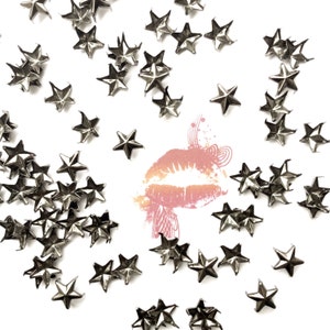 May include: A white background with a scattering of silver star-shaped metal studs. The studs are arranged in a loose pattern, with a few clustered together and others spread out. There is a pink and orange lip print in the center of the image.