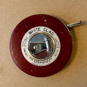May include: A vintage red and silver Lufkin White Clad steel measuring tape. The tape measure has a circular casing with the text "WHITE CLAD" and "50 FT." The casing is marked "THE LUFKIN RULE CO. SAGINAW, MICH. U.S.A."