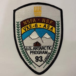 May include: Embroidered patch with a shield shape, featuring text and graphics related to the U.S. Antarctic Program. The patch includes the text "NSFA NSF VXE-6 ASA" and "U.S. ANTARCTIC PROGRAM 93". The design incorporates mountains, a building, and a laurel wreath.