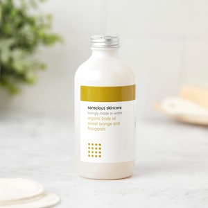 May include: A white bottle with a silver cap, labeled "conscious skincare" and "organic body oil sweet orange and frangipani." The label has a gold band and a dotted design. The bottle is likely filled with body oil.