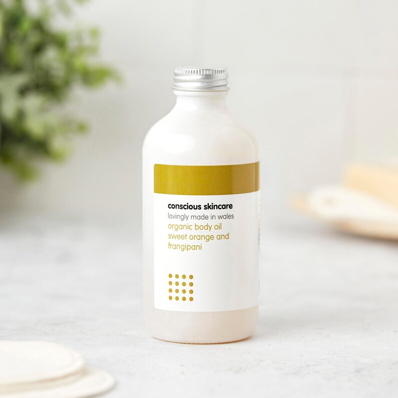May include: A white bottle with a silver cap, labeled "conscious skincare" and "organic body oil sweet orange and frangipani." The label has a gold band and a dotted design. The bottle is likely filled with body oil.