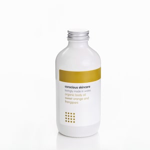 May include: A white bottle with a silver cap and a gold label. The label reads "conscious skincare" and lists the product as organic body oil with sweet orange and frangipani. The bottle is on a white background.