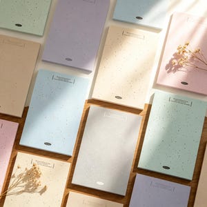 May include: A collection of pastel-colored notepads with speckled patterns. Each notepad features a rectangular design with rounded corners and a small printed logo. The notepads are arranged on a wooden surface, with some accented by dried flowers.