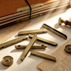Op de afbeelding: Een verzameling antiek messingkleurige metalen paginahouders en decoratieve elementen. De afbeelding toont een leren gebonden notitieboek, een waszegelstempel en verschillende metalen clips, wat een vintage of stationery thema suggereert.