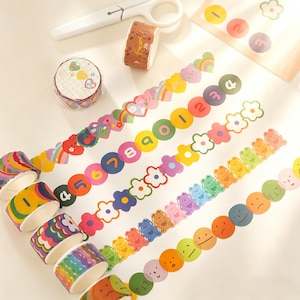 Puede incluir: Una colección de cintas washi decorativas con varios diseños, incluyendo corazones, arcoíris, números, flores, osos y caras sonrientes. Se ven varios rollos de cinta, junto con tiras de cinta decorativa y unas tijeras blancas.