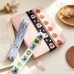 May include: A collection of decorative ribbons with various patterns, including cartoon faces, stars, and fruit. The ribbons are in shades of blue, black, and white, and are placed on a pink notebook. A few ribbons are visible.