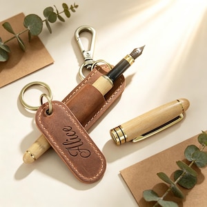 May include: A wooden fountain pen with gold accents and a black nib, alongside a matching pen holder and keychain. The keychain has a leather tag with the name "Alice" engraved. The pen is displayed with a brown leather pen case.