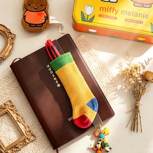 May include: A brown leather journal with a colorful sock-shaped pen holder. The pen holder is yellow with green, blue, and red accents. A yellow tin with the text "miffy melanie" is in the background, along with a small plush toy and a charm.