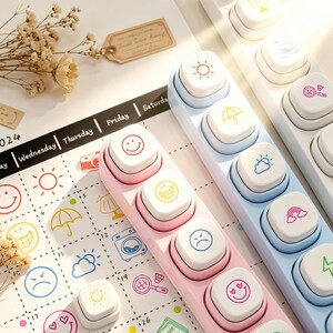 Könnte beinhalten: Nahaufnahme von drei Reihen von Stempeln zum Thema Stimmung und Wetter in Rosa, Blau und Weiß. Jeder Stempel hat eine quadratische weiße Oberseite mit einem anderen Symbol oder Emoji. Im Hintergrund befindet sich ein Kalender mit passenden Symbolen.